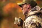 Caucasian Hunter in His 40s Wearing Camouflage Drinking a Coffee. Hunting Season Theme. Visual concept for a blog discussing hunting accidents and liability.